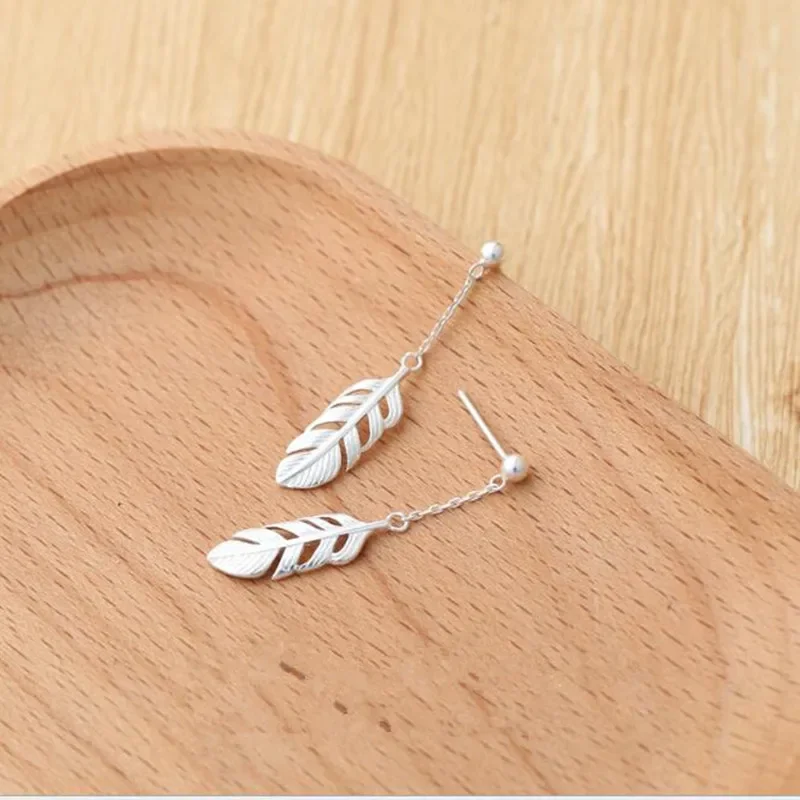 A pair of silver-toned metal feather drop earrings with thin chains on a wooden tray.