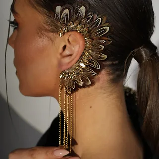 A model wearing an arch-shaped gold-toned ear cuff with feather-shaped detailing, faceted accents, and fine hanging chains.