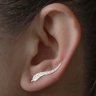 A close-up of a gold-toned ear crawler shaped like a feather, worn along the curve of a human ear.