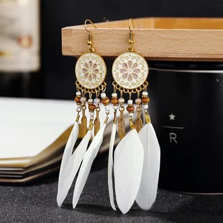 A pair of drop earrings with a white floral-patterned metal disc top, beaded chains, and white feathers hanging from a wooden stand.