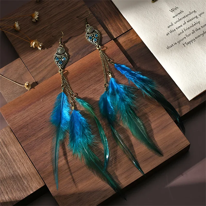 Two blue and green gradient feather drop earrings with antiqued metal shield tops and chain details resting on a wooden surface.