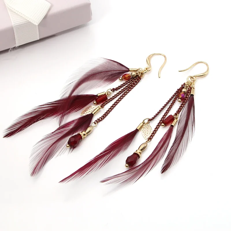 A pair of deep red feather earrings with hanging chains, beads, and small gold-toned leaf charms.