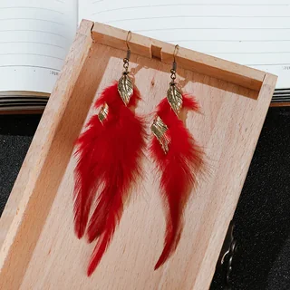 Bright red feather drop earrings with gold-tone wing-shaped charms resting on a wooden tray.