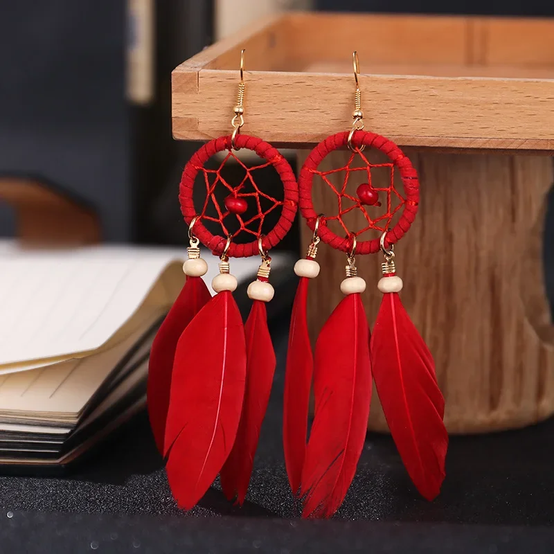 A pair of red dreamcatcher-style drop earrings featuring a circular webbed frame with hanging feathers and small beads.