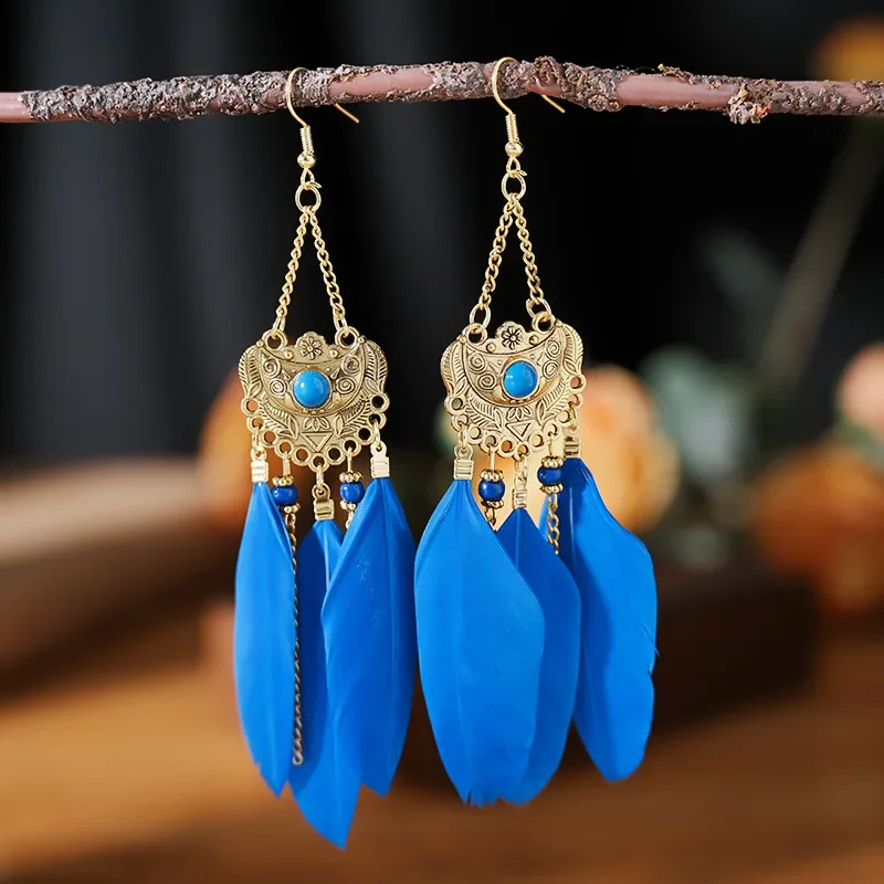 Two drop earrings featuring ornate stamped metal discs, central stone accents, hanging chains, and bright blue or red plumes.