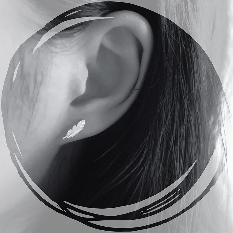A close-up of a silver-toned feather-shaped stud earring worn on an earlobe, showing fine etching and a curved shape.