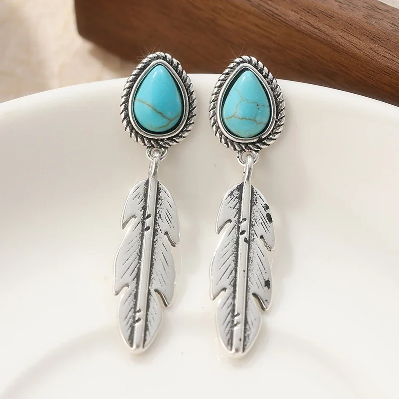 A pair of drop earrings with a turquoise-colored teardrop stone and a hanging silver-toned metal feather.
